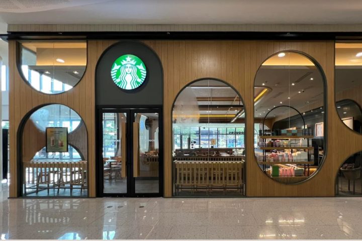 モール内にあるスターバックスの外観。木目調の壁に丸い窓が並び、ガラス越しに店内のテーブル席やカウンターが見える。正面入口上部に緑色のスターバックスロゴが掲げられている。